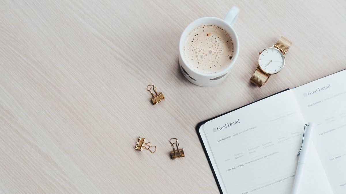 A minimalist desk arrangement featuring a coffee cup, notebook, pen, and wristwatch.